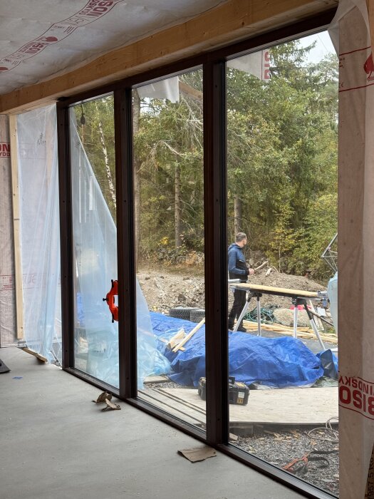 Bilden visar ett pågående byggprojekt genom glasrutor, med en person utomhus vid en arbetsbänk omgiven av byggmaterial och täckplast.