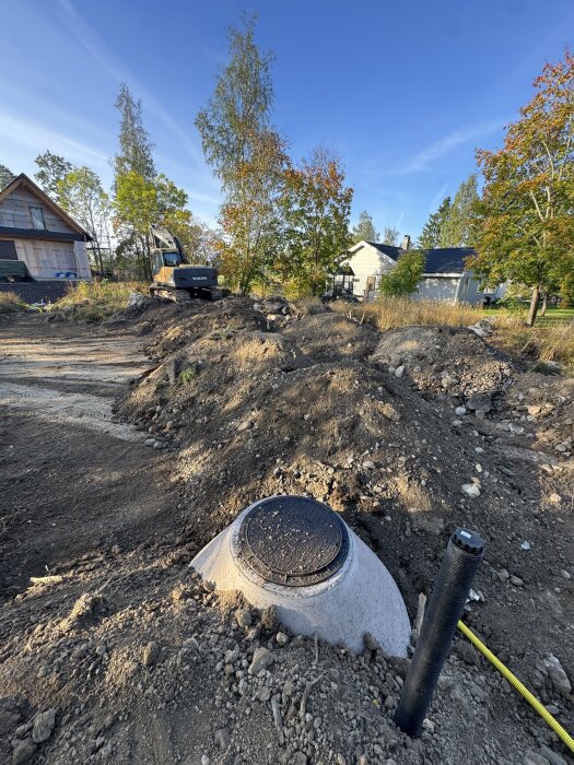 Tomt med grävmaskin och jordhögar, avloppslock och rör synliga. Hus i bakgrunden, omgivna av träd, under klar himmel.