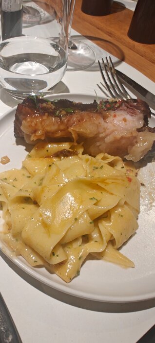 Tallrik med pappardellepasta i krämig sås och köttbit toppad med örter. Bakgrund med glas och bestick.
