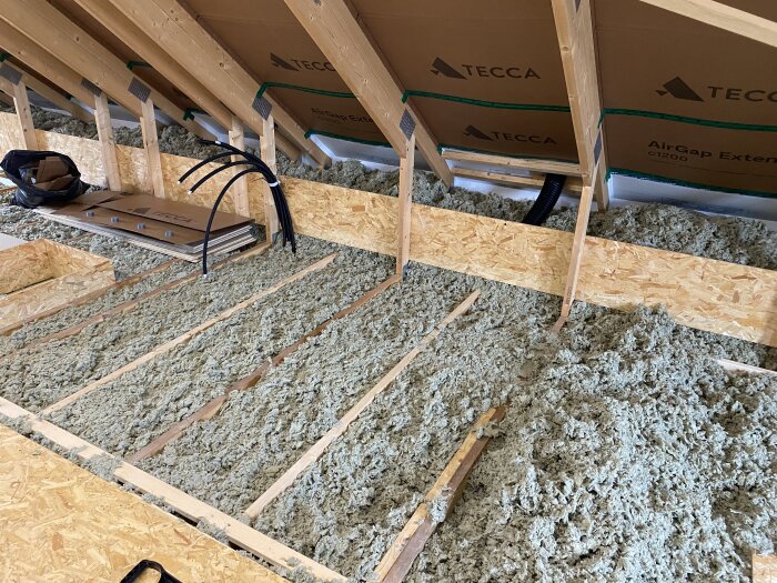 Oinredd övervåning med isolering på golvet, synliga takstolar, ventilationsrör och byggmaterial.