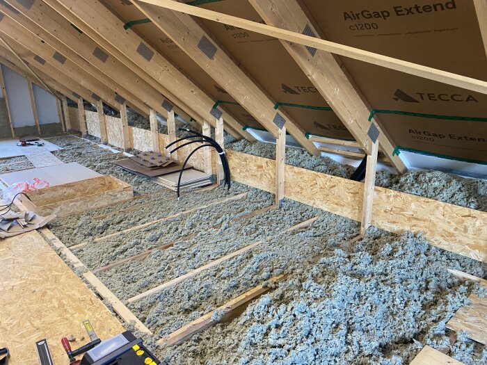 Oinredd övervåning med isolering mellan takstolar, synliga ventilationsrör och byggmaterial. Väggar klädda med luftspaltskivor.