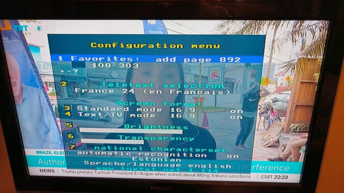 TV-skärm visar teletext-konfigurationsmeny med val som "Teletext selection", "Screen format" och "Brightness". Bakgrund med utomhusscen.