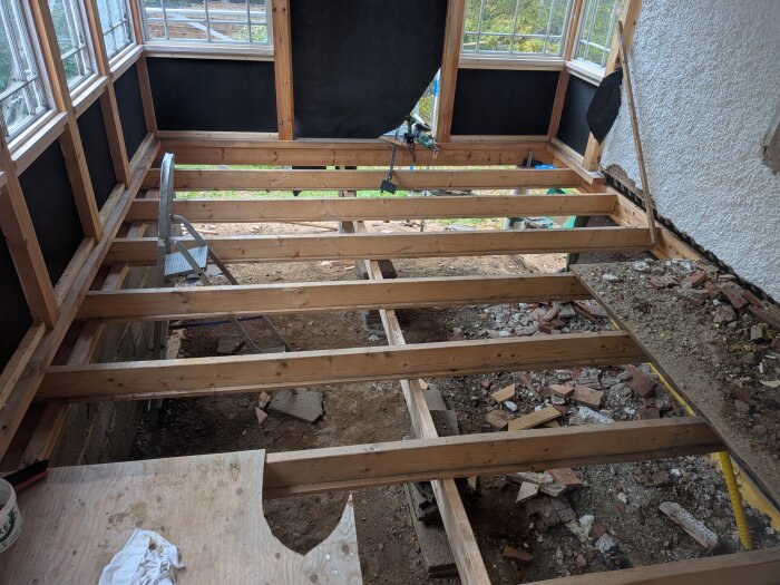 Träkonstruktion av ett golv under uppbyggnad i en glasveranda med bjälkar synliga och tegelrörelser på marken. Stege och fönster syns i bakgrunden.
