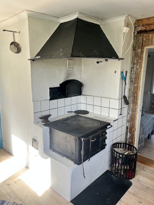 Vedspis och bakugn med svart huva och två luckor i ett kök, omgiven av vita kakelplattor, med vedkorg och vägg i trä.