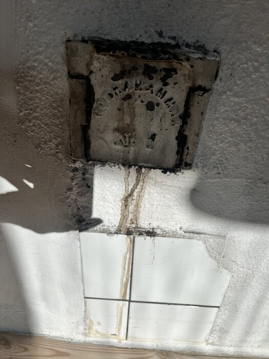 Inmålad lucka på en vedspis med brunaktiga rännor på väggen under, nära kakelplattor.