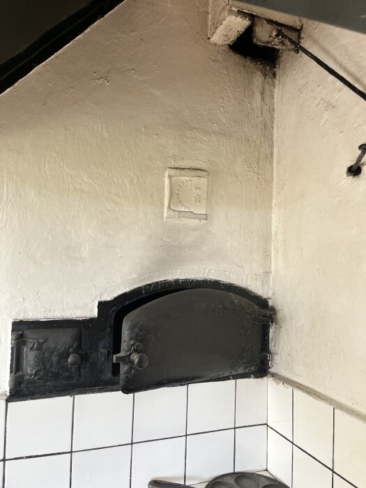 Svart vedspis och bakugn med två luckor mot vitkaklad vägg, delvis öppnade spjäll och tecken på tidigare fuktinträngning.