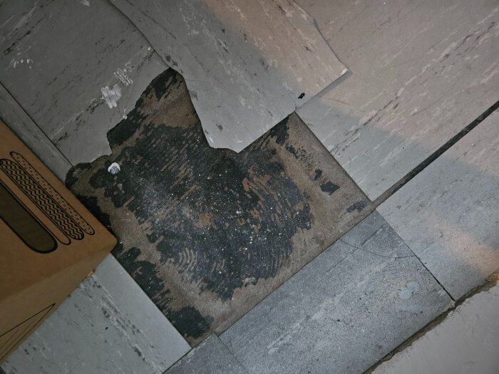 Skadat golv med lösa, grå plattor och ett mörkt område synligt under. En kartongkant syns till vänster i bilden.