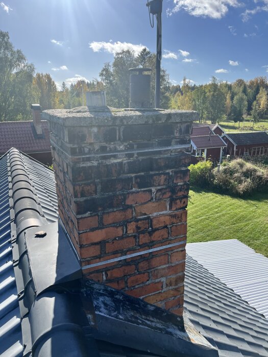 Tegelstensskorsten på tak med omgivande träd och ängar i bakgrunden, fotograferad i dagsljus.