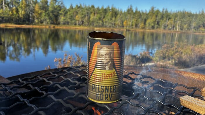 Konservburk med Bullens pilsnerkorv värms på en grill vid en sjö med skog i bakgrunden.