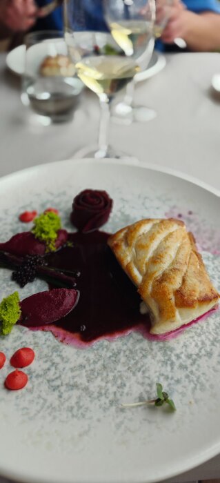 En elegant tallrik med grillad fisk, rödbetssås och dekorationer, samt ett glas vitt vin i bakgrunden på en lunchrestaurang i hamnen.