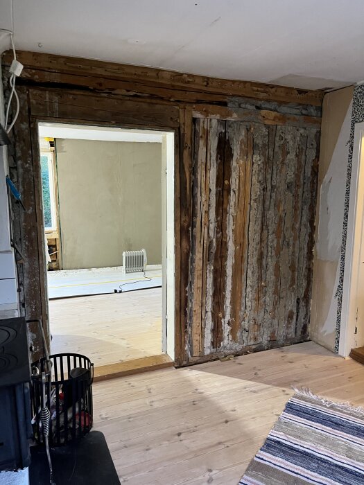 Gammal vägg i hus under renovering med delvis avlägsnade ytskikt och synlig stomme, samt öppning till angränsande rum med ljus parkett.