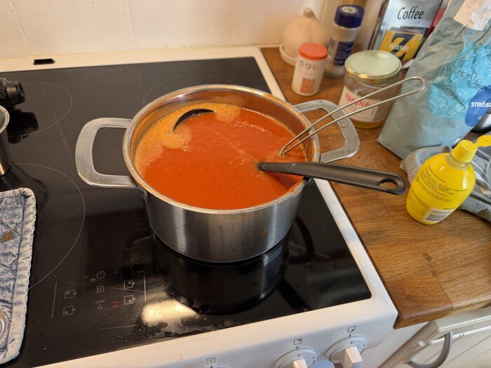 Kastrull med tomatsoppa på en spishäll, omgiven av köksartiklar som citronjuiceflaska, sockerpåse och kryddburkar.