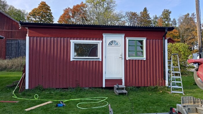 Rött hus med ny panel och vita fönster, stege lutad mot väggen. Hemgjort renoveringsprojekt med fokus på takhöjning och fönstermontering.
