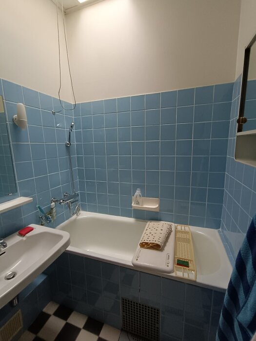 Badrum med blå kaklade väggar, ett badkar med tillbehör, handfat och hyllor, svartvitt rutigt golv.