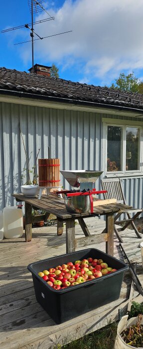 Svart plastlåda full med röda och gröna äpplen står på en träveranda med en äppelpress och kvarn bredvid.