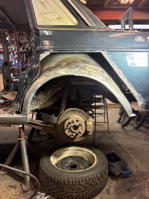 Bilens bakre hjulområde på en domkraft i ett garage, utan hjul monterat, med en rostig bromsskiva och ett hjul på golvet bredvid.
