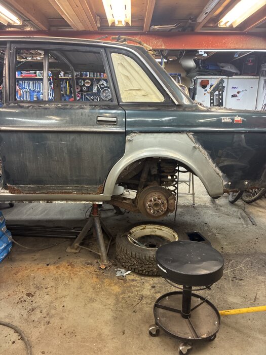 En bil på pallbockar i en verkstad med avtagna hjul, verktyg i bakgrunden och ett garagegolv med rostfläckar.