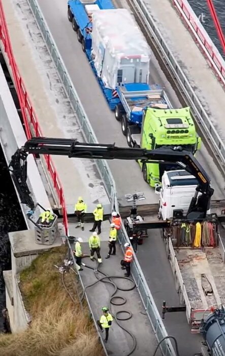 En grupp arbetare inspekterar och utför arbete på en bro. En kran är synlig, tillsammans med en lastbil och säkerhetsutrustning.