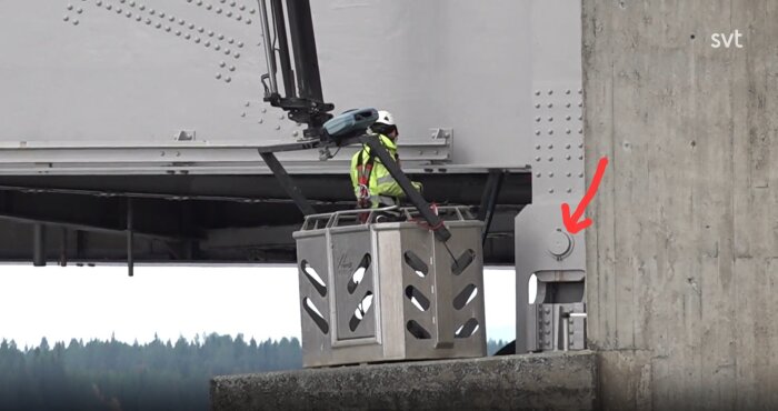 Byggarbetare i säkerhetsutrustning arbetar från en lyftkorg nära en brokonstruktion, med en röd pil som pekar på en bult på bron.