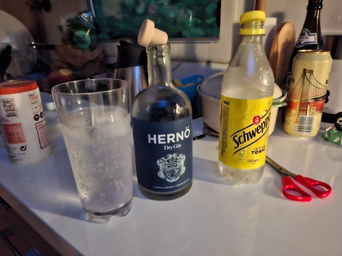 Flaska med Hernö dry gin, en Schweppes tonic-flaska och ett glas med dryck på en köksbänk, omgiven av hushållsartiklar och röda saxar.