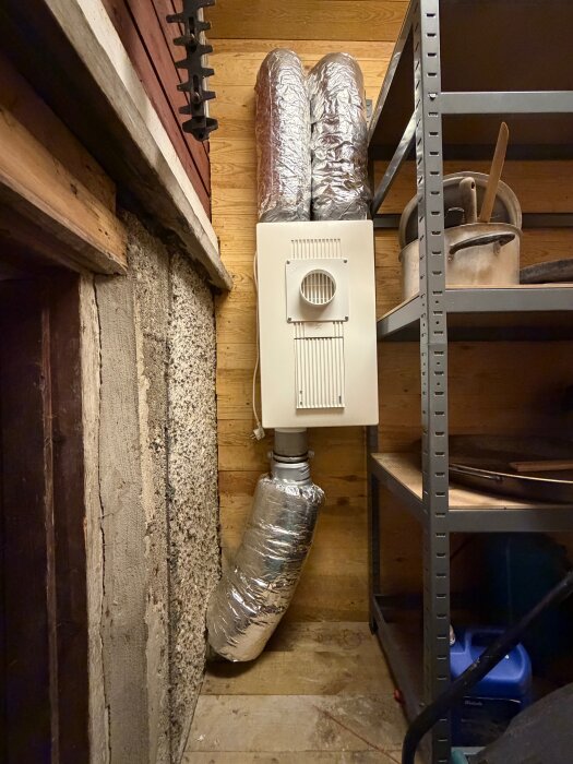 Ventilationssystem med silverfärgade isolerade rör i ett träpanelrum, bredvid en metallhylla med köksartiklar.