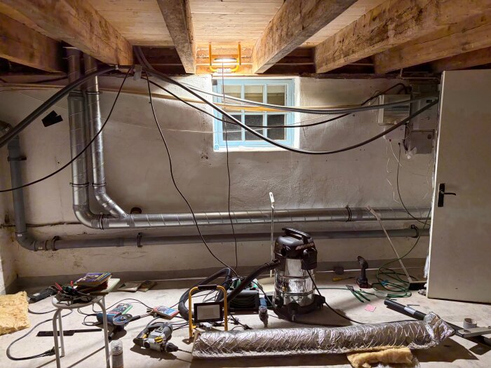 Källare under renovering med synliga rör, verktyg och kablar på golvet, inspektionsfönster och arbetslampa upplyser rummet.
