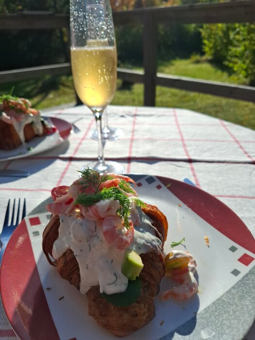 Croissant med räkfyllning och dill på en tallrik, bredvid ett glas champagne, står på ett bord utomhus med en rutig duk.