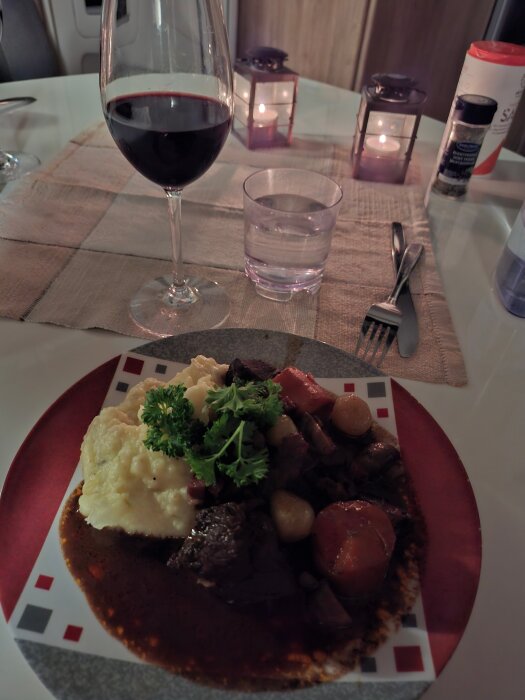 Boeuf bourguignon med potatis- och jordärtskockstomp på en tallrik, uppdukat med ett glas rödvin och ljuslyktor i bakgrunden.