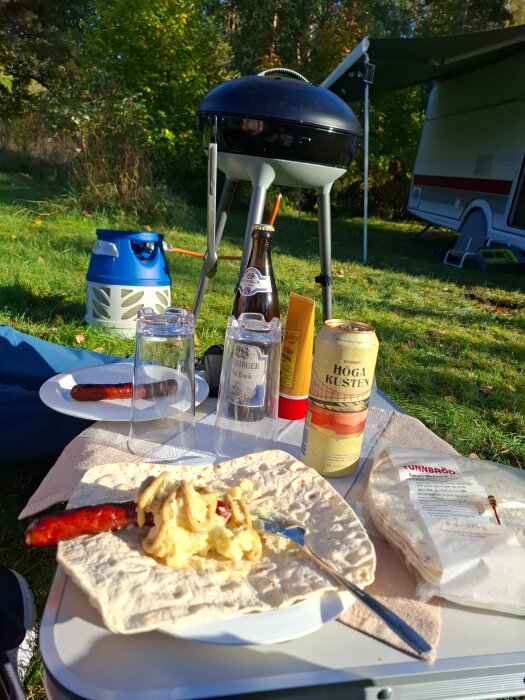 Korv med tunnbröd och potatissallad på campingbord, grill i bakgrunden, omgiven av natur och husvagn.