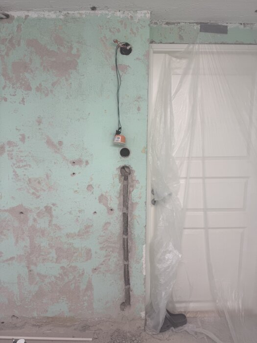 Vägg under renovering med löst sittande dosor och elrör, plastering skyddar dörr. Spår för elinstallation synliga.