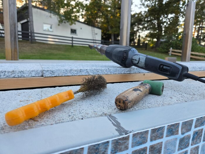 Verktyg vid pool, lödkolv, pensel och rengöringsverktyg på poolkant med trä och mosaik i bakgrunden.