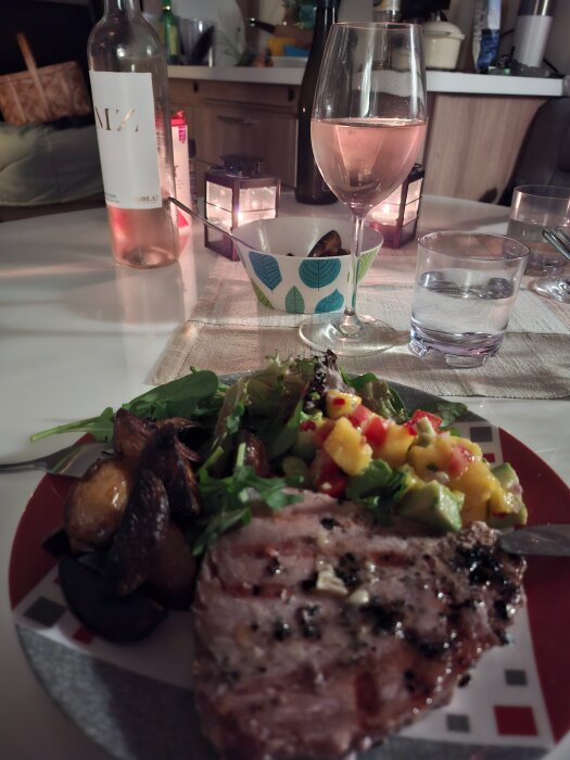 Tallrik med grillat kött, potatis, sallad, mango- och avokadosalsa; vinflaska, vinglas och ljus på bordet i bakgrunden.