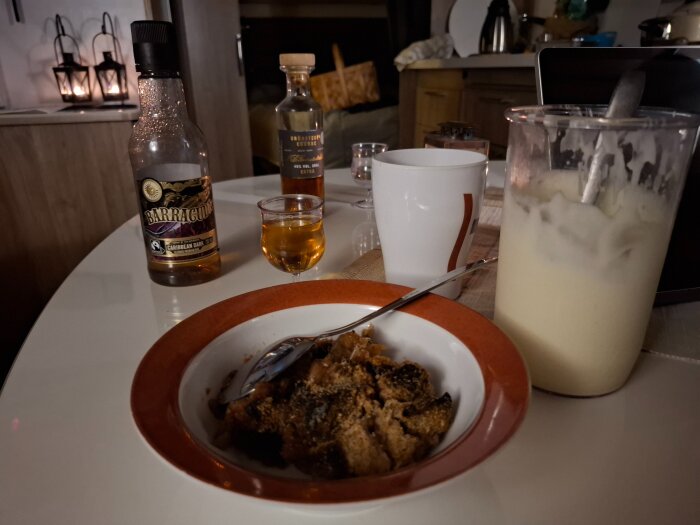 En dessert med grädde i en skål på ett bord, omgiven av en flaska sprit och en mugg, med tända ljus i bakgrunden.