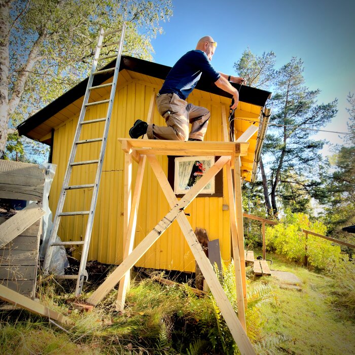 Person bygger tak på gult hus, står på träställning. Stege lutar mot väggen. Soligt, grönskande omgivning.