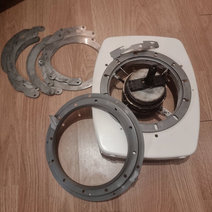 Demonterad köksfläkt på trägolv med lösa metalldelar och en plastbas visar olika komponenter, bland annat ett spjäll och metallringar.