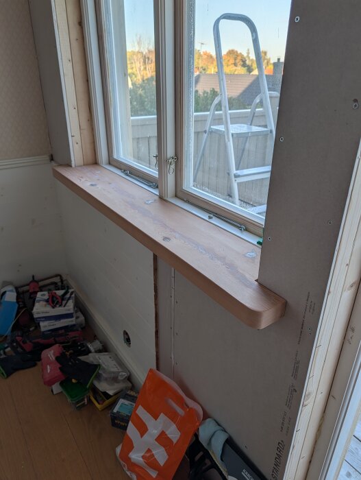 Fönsterbräda av återvunna plank, med utsikt över trädgård och en stege utanför; verktyg och material på golvet.