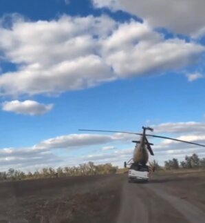 En helikopter transporteras ovanpå en lastbil på en landsväg under en blå himmel med moln.