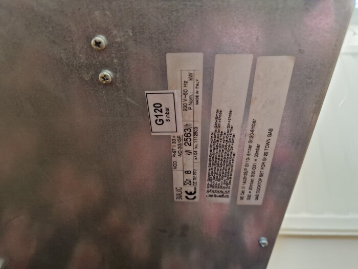 Närbild av en metallplatta med etiketter och skruvar, visar specifikationer för en gasanordning; modellnummer och tech-info synligt.