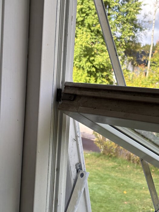 Öppet fönster med saknat plastbeslag, visar detaljer av fönsterkarm och trädgård i bakgrunden.
