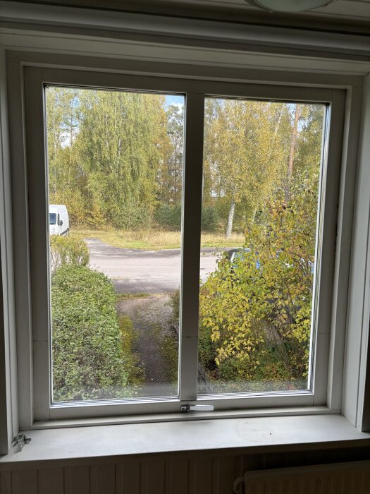 Utsikt genom fönster mot trädgårdsstig, gröna buskar och höstklädda träd; en vit skåpbil står parkerad till vänster.