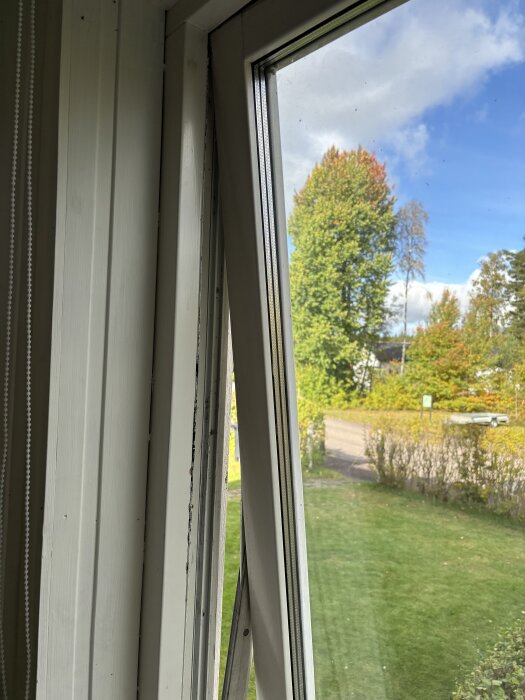 Öppet fönster inifrån sett, med utsikt över trädgård och träd, en fönsterkarm saknas på insidan.