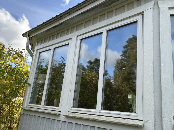 Ett vitt trähus med stora fönster som reflekterar träd och en blå himmel med moln.