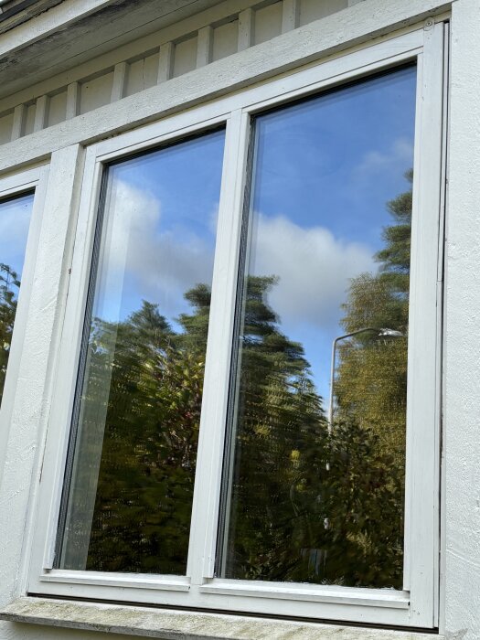 Fönster med vit träram som reflekterar träd och blå himmel med moln.