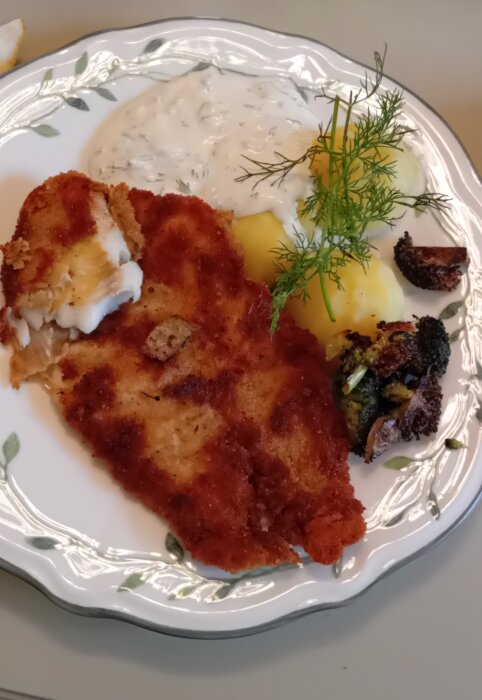 Pankopanerad fisk med citron och gräslökssås, ångad broccoli och potatis toppad med dill på en tallrik.