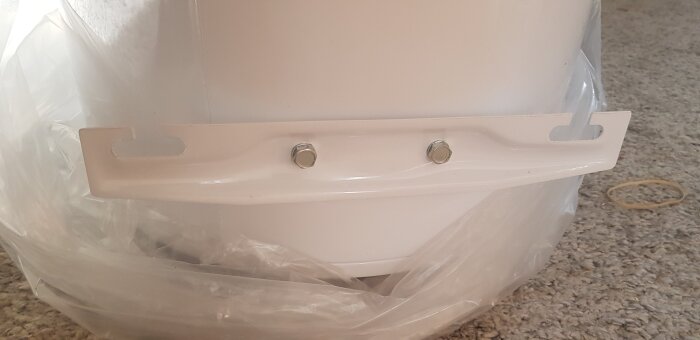 Detalj av en fastsättningsskena med två skruvar, troligen för en varmvattenberedare, placerad på en matta med plastskydd.