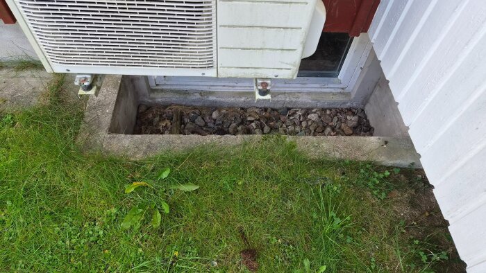 Betongrännor fyllda med grus vid husvägg, gräs runtomkring och ett yttre element monterat ovanför på väggen.