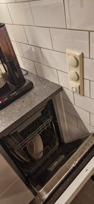 Öppen diskmaskin vid köksdisk och väggen med dimmer och golvtermostat synliga ovanför.
