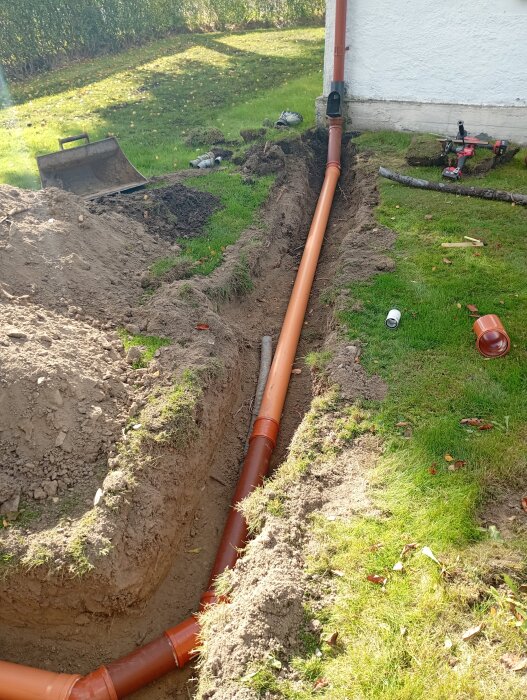 Orange dräneringsrör installerat i en grävd trädgårdsgrop nära ett hus, med verktyg och jord runtomkring.