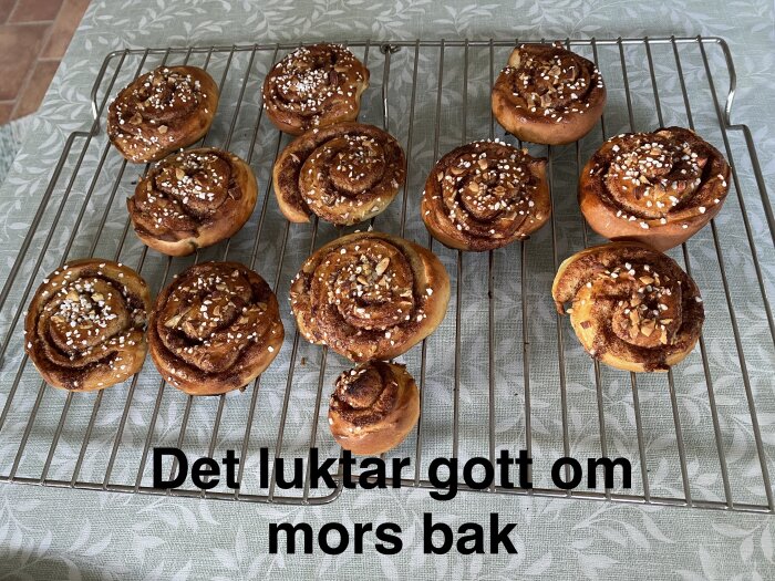 Kanelbullar med pärlsocker och nötter ligger på ett galler på en blommig duk. Texten "Det luktar gott om mors bak" syns nedanför.