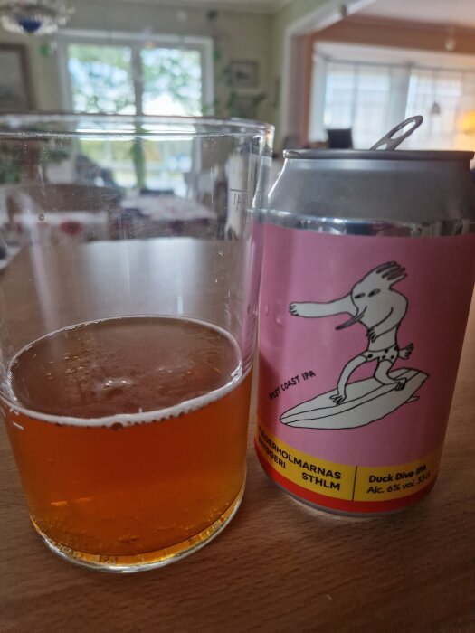 Burk med Fjäderholmarnas Duck Dive IPA och ett glas med öl på ett bord i ett rum med fönster i bakgrunden.